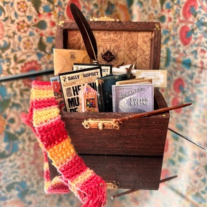 Op de afbeelding: Een houten kist gevuld met miniatuurboeken, een ganzenveer en een rood-gele sjaal. De kist is open en de boeken zijn zichtbaar. De kist staat op een gepatroneerd oppervlak.