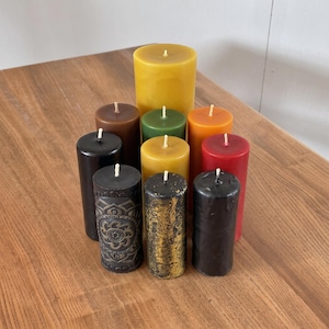 Puede incluir: Una colección de diez velas cilíndricas de varios colores, incluyendo amarillo, verde, naranja, rojo, marrón y negro. Algunas velas tienen superficies lisas, mientras que otras muestran intrincados patrones en relieve o acabados dorados.