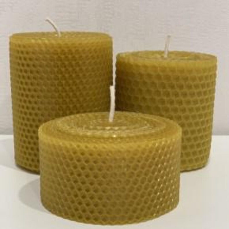 May include: Three cylindrical beeswax candles with a distinct hexagonal honeycomb texture on the exterior. Each candle features a single white cotton wick centered at the top. The candles are golden yellow in color and vary in height.