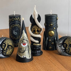 Puede incluir: Una colección de seis velas decorativas de diversas formas. Dos son redondas con caras de sol y luna, una es un cilindro negro con un símbolo yin-yang, una es un cono con un ojo, una presenta un árbol y una serpiente, y otra muestra planetas.