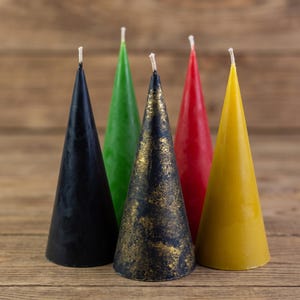 Puede incluir: Un conjunto de cinco velas en forma de cono dispuestas en grupo. Las velas presentan colores sólidos que incluyen negro, verde, rojo y amarillo, con una vela negra central decorada con un acabado metálico dorado texturizado y una mecha.