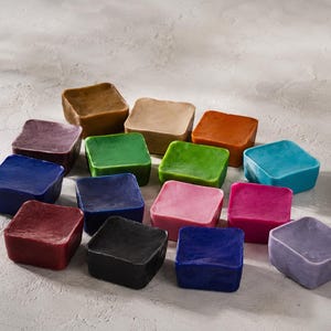 Puede incluir: Una colección de quince bloques de cera cuadrados en una variedad de colores sólidos que incluyen marrón, beige, naranja, verde, verde lima, azul verdoso, morado, azul, azul oscuro, rosa, magenta, granate, negro, azul marino y lavanda, dispuestos.