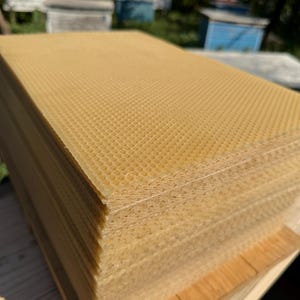 May include: A stack of rectangular beeswax foundation sheets featuring a consistent hexagonal honeycomb pattern. The sheets are a translucent golden yellow color, showing a textured grid structure on the surface and layered edges.