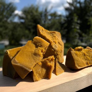 May include: Several large, irregular chunks of raw, golden-yellow beeswax with a dense, solid texture. The pieces display a natural, matte finish with visible variations in color and surface grain, arranged in a cluster on a light-colored wooden surface.