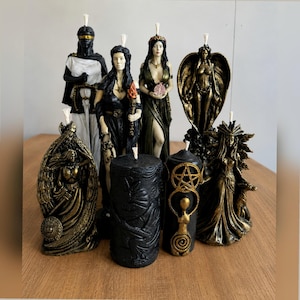 Puede incluir: Una colección de ocho velas figurativas que representan diversas deidades mitológicas y motivos simbólicos. Las velas tienen acabados en negro, dorado y blanco, con figuras aladas, una antorcha, una granada y un pentagrama entre los diseños.