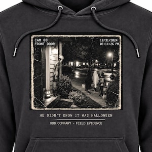 May include: A charcoal gray hooded sweatshirt featuring a graphic print of a security camera still showing an alien and children in costumes on a porch. The text reads CAM 03 FRONT DOOR, 10/31/2024, 08:14:26 PM, HE DIDN'T KNOW IT WAS HALLOWEEN, ODD COMPANY - FIELD EVIDENCE.