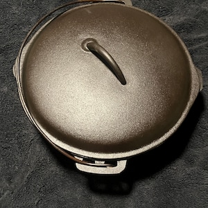 May include: A round, dark gray cast iron Dutch oven with a matching lid. The lid features a central curved handle, and the pot includes a thin, arched metal bail handle attached to two side lugs. The surface has a textured, matte finish.