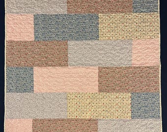 May include: A rectangular quilt featuring a brick-patterned layout of alternating fabric blocks. The design includes various floral prints in shades of blue, pink, cream, and brown, finished with a light-colored binding along the outer edges.