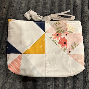 May include: A rectangular fabric pouch featuring a patchwork design of white, mustard yellow, navy blue, and coral pink triangles. The pouch includes a floral print section and a light gray fabric tie closure at the top edge.