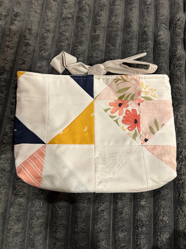 May include: A rectangular fabric pouch featuring a patchwork design of white, mustard yellow, navy blue, and coral pink triangles. The pouch includes a floral print section and a light gray fabric tie closure at the top edge.
