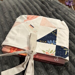 May include: A quilted fabric book sleeve featuring a patchwork design of white, soft pink, and dark navy blue triangles with floral and script patterns. The sleeve is tied with a light beige fabric ribbon and holds a book titled The House of Eve.