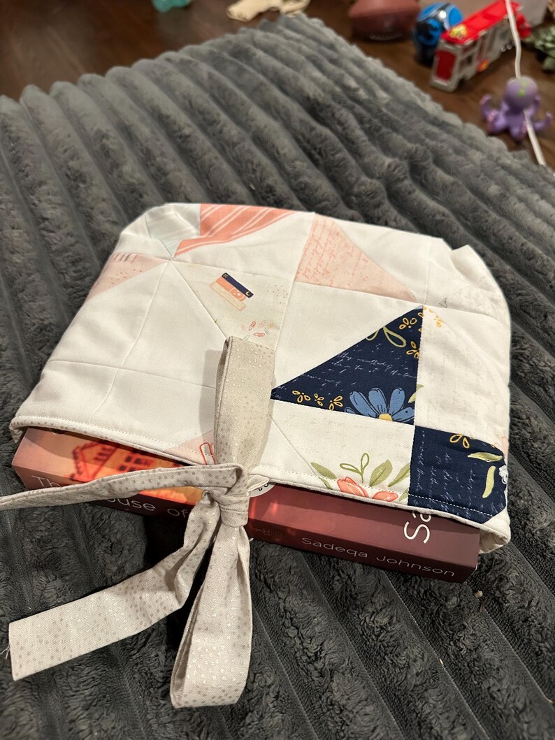 May include: A quilted fabric book sleeve featuring a patchwork design of white, soft pink, and dark navy blue triangles with floral and script patterns. The sleeve is tied with a light beige fabric ribbon and holds a book titled The House of Eve.