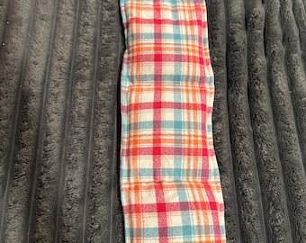 May include: A long, rectangular fabric heating pad featuring a plaid pattern in shades of red, orange, and light blue on a cream-colored background. The item has stitched seams creating multiple sections along its length.