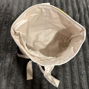 May include: A fabric bucket-style bag with an open top, featuring a light beige interior lining patterned with small, scattered white dots. The exterior shows a glimpse of a bright yellow fabric section, and two fabric ties hang from the base.