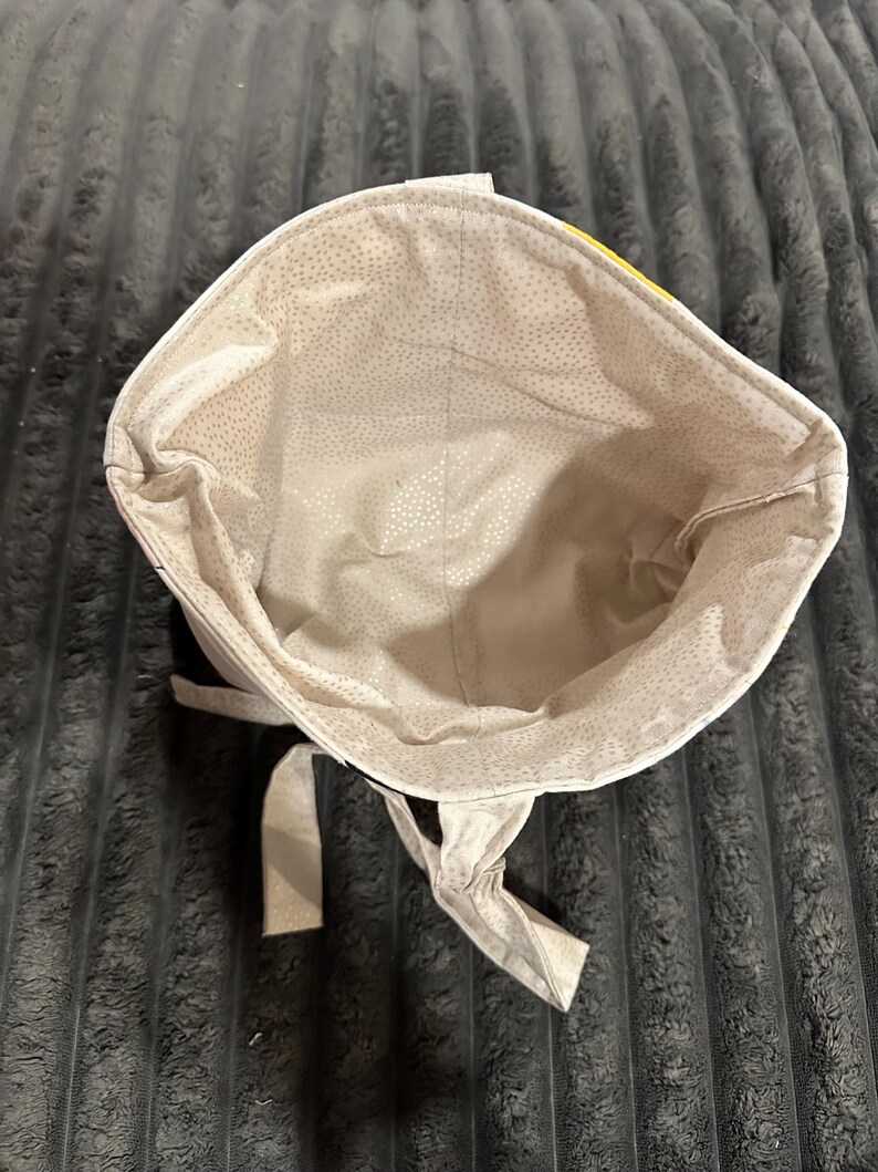 May include: A fabric bucket-style bag with an open top, featuring a light beige interior lining patterned with small, scattered white dots. The exterior shows a glimpse of a bright yellow fabric section, and two fabric ties hang from the base.