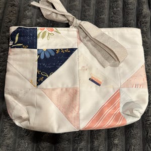 May include: A quilted fabric pouch featuring a patchwork design of white, navy blue, and soft pink panels. The fabric includes floral patterns, script text, and small book illustrations. A light gray tie closure is attached to the top edge.
