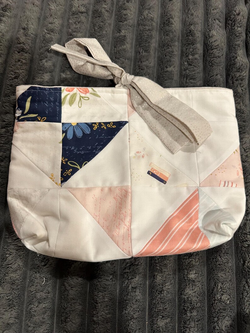 May include: A quilted fabric pouch featuring a patchwork design of white, navy blue, and soft pink panels. The fabric includes floral patterns, script text, and small book illustrations. A light gray tie closure is attached to the top edge.