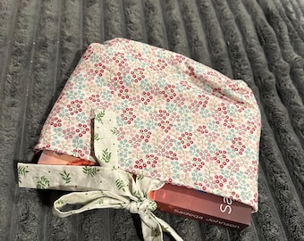 May include: A fabric book sleeve featuring a small floral pattern in pink, red, and blue on a white background. The sleeve has a tie closure made of a light-colored fabric with a green leaf print, partially covering a book by Sadeqa Johnson.