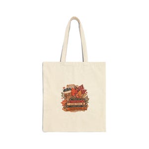 May include: A beige canvas tote bag featuring a central graphic of a stack of books, a coffee cup, and autumn leaves. The book spines are labelled with the text Crisp Air, Cozy Days, Autumn Stories, Pumpkin Spice, and Sweater Weather.