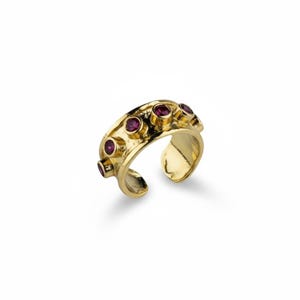 Puede incluir: Un anillo abierto de tono dorado con superficie texturizada y cinco piedras pequeñas, redondas y de color rosa intenso, engastadas individualmente a lo largo del borde superior. El anillo tiene un interior ancho y pulido con abertura trasera.
