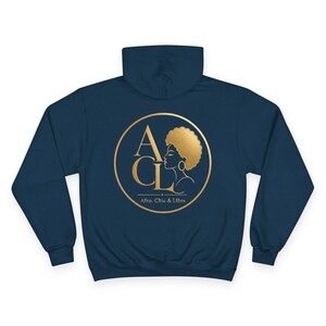 Könnte beinhalten: Ein dunkelblauer Kapuzenpullover mit einer goldenen kreisförmigen Grafik auf der Rückseite. Das Design enthält die Buchstaben ACL, das Profil einer Person mit Afro-Frisur und den Schriftzug Afro, Chic & Libre in goldener Farbe.