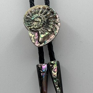 May include: A bolo tie featuring a central iridescent ammonite fossil pendant with swirling patterns in shades of green, purple, and gold. The pendant is attached to a black braided cord, which terminates in two matching iridescent shell tips.