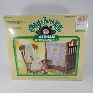 May include: A Cabbage Patch Kids Afghan Stenciling Kit box, number 26251, featuring a yellow and white color scheme. The packaging displays a wooden chair with a stenciled blanket, a crib with a matching blanket, and a doll. Text includes kit contents.