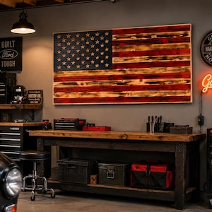 May include: A rectangular wooden wall art piece featuring a charred American flag design with a dark blue canton containing white stars and horizontal stripes in alternating burnt red and natural wood tones, mounted on a gray wall in a garage.
