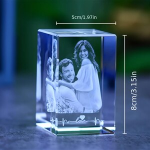Puede incluir: Un bloque de cristal transparente rectangular con una imagen grabada en láser 3D de una pareja abrazada. La base incluye un gráfico de línea de latido con la palabra "love" y un símbolo de corazón. Mide 5 cm de ancho por 8 cm de alto.