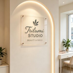 May include: A clear rectangular acrylic sign mounted on a wall with four metal standoffs. The sign features a black logo with a leaf icon above the text Trilumi STUDIO BEAUTY & NAILS in a combination of script and sans-serif black lettering.