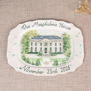 Pode incluir: Uma travessa oval de cerâmica branca com uma ilustração pintada à mão de uma casa grande de dois andares com telhado azul, rodeada por árvores verdes. O texto "The Magdalene House" e "November 23rd 2026" está escrito em cinza escuro.