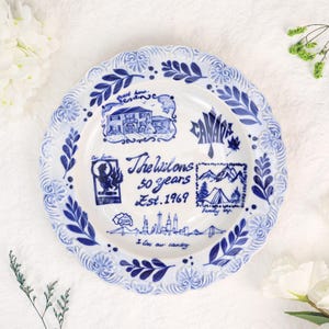 May include: A white ceramic plate with a blue decorative border and blue illustrations. The centre features the text "The Wilsons 50 years Est. 1969" surrounded by drawings of a house, a maple leaf, mountains, a tent, and a city skyline.