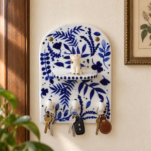 May include: A white ceramic wall-mounted key holder with a blue botanical pattern, featuring an arched top, a small central shelf, and three hooks. A small, painted ceramic cat figurine sits on the shelf, and three sets of keys hang from the hooks.