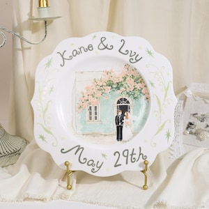 Puede incluir: Un plato decorativo blanco de bordes festoneados con una ilustración pintada a mano de una pareja de boda frente a un edificio azul verdoso con flores rosas. El texto dice Kane & Ivy y May 29th en gris con detalles verdes.