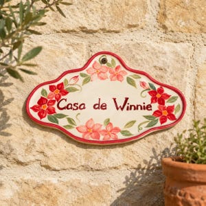 Pode incluir: Uma placa de parede de cerâmica de formato oval com uma borda vermelha recortada, apresentando flores vermelhas e folhas verdes pintadas à mão sobre um fundo creme. O texto "Casa de Winnie" está escrito ao centro em fonte cursiva vermelha.