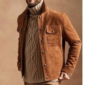 May include: A tan suede jacket featuring a structured collar, long sleeves, and two chest pockets with button closures. The jacket is worn over a beige cable-knit sweater with a high turtleneck, showcasing a textured vertical pattern.