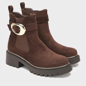 May include: A pair of dark brown suede ankle boots featuring a thick lug sole, a side strap with a large polished gold-tone oval buckle, and a pull tab at the heel. The boots have a rounded toe and a dark brown elastic side panel.