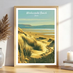 Puede incluir: Una lámina artística enmarcada con una ilustración estilizada de Woolacombe Beach, Devon. La imagen muestra dunas de arena dorada con hierbas altas en primer plano, que conducen a una playa ancha, colinas distantes y un cielo azul. El texto dice Woolacombe Beach Devon.