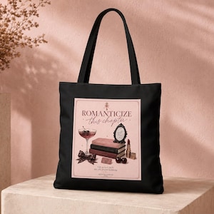 Può includere: Una borsa tote nera con una grafica rettangolare sul davanti. La grafica ha uno sfondo rosa chiaro con il testo ROMANTICIZE this chapter e fall in love with the life you're building. Raffigura un bicchiere da cocktail, libri, uno specchio, un rossetto e un fiocco.