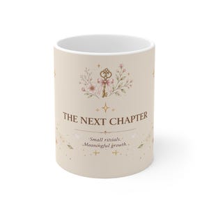Può includere: Una tazza in ceramica beige con un'illustrazione di una chiave dorata con un fiocco rosa, accenti floreali e il testo THE NEXT CHAPTER seguito da Small rittials. Meaningful growth. Il design include piccole stelle e scintille dorate.