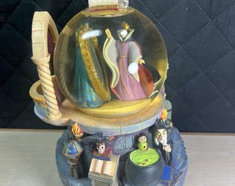 May include: A Disney snow globe featuring the Evil Queen and a magic mirror inside the glass sphere. The base depicts a stone castle scene with a black cauldron, a green potion, a skull, books, and a raven. The queen wears a purple and black gown with a gold crown, while the mirror has a gold frame.