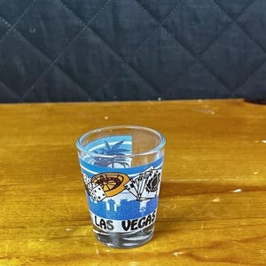 May include: A clear glass shot glass featuring a blue graphic print of Las Vegas, including a palm tree, a roulette wheel, dice, playing cards, and the text LAS VEGAS in black lettering around the base of the glass.