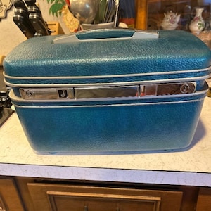 May include: A teal blue hardshell train case with a textured exterior, a silver-toned handle, and a central metal latch plate featuring the letter J. The rectangular vanity case has a structured design with silver trim accents along the middle.