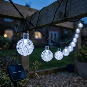 May include: A string of outdoor solar-powered LED lights featuring spherical bulbs with a cracked glass texture. The clear, round globes emit a bright white light and are connected by a black wire, accompanied by a small black solar panel stake.