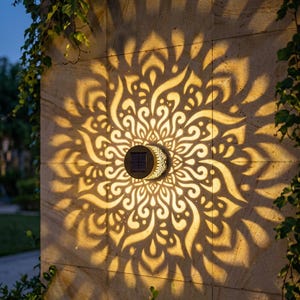 May include: A circular solar-powered wall light with a dark centre and a metallic outer casing, mounted on a stone surface. The light projects a large, intricate, golden-yellow mandala-style pattern of swirling shapes and lines onto the wall.