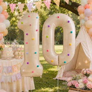 May include: Two large cream-colored number balloons shaped as a 1 and a 0, decorated with small, colorful pom-poms in shades of pink, blue, yellow, and purple. The balloons are suspended in an outdoor setting, featuring a festive party display.