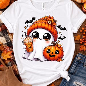 Può includere: Una t-shirt bianca con la grafica di un simpatico fantasma che indossa un berretto di lana arancione con fiori, tiene in mano un caffè freddo con un disegno di zucca e si trova accanto a una zucca sorridente con tre pipistrelli neri.