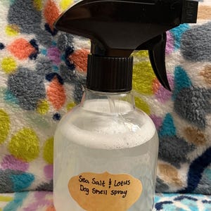 Op de afbeelding: Een doorzichtige plastic spuitfles met een zwarte sproeikop die een doorschijnende vloeistof bevat. Op de voorkant zit een beige etiket met een geschulpte rand en de handgeschreven tekst: Sea Salt & Lotus Dog Smell spray in zwarte inkt.