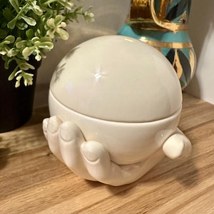 Può includere: Un contenitore in ceramica color crema a forma di mani giunte che sostengono un coperchio sferico liscio. L'oggetto presenta una finitura lucida e un design arrotondato, con le dita e i palmi che formano la base per sostenere il coperchio.