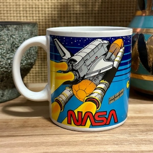 Peut inclure: Un mug en céramique blanche orné d'un graphisme coloré représentant une navette spatiale de la NASA en plein décollage. Le motif comprend la navette, des flammes d'échappement jaune vif, un satellite et le logo rouge NASA en bas.